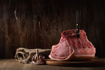 a whole freshly cut piece of raw pork loin lies on a cutting board with a knife, burlap and garlic a brown wooden background. side view. artistic moody photo in simple rustic style with copy space