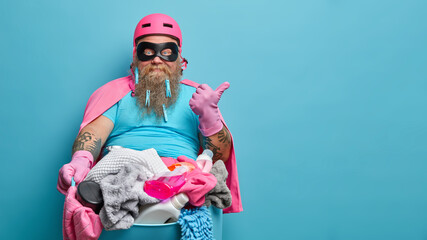 Busy bearded man with thick beard does laundry at home wears eyemask helmet and cloak points thumb...