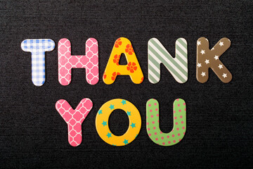 Card with Thank You words made from mixed vivid colored wooden letters on a textured dark black textile material that can be used as a message.