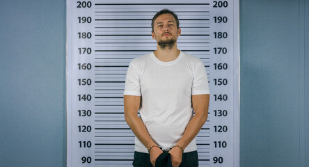 Arrested a man in handcuffs standing near a white board with a scale. Mugshot