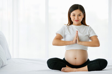 young pretty Asian woman sitting on a bed joins her hands in front of her chest with a smiley face...
