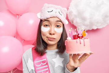 Close up portrait of good looking young Asian female model applies patches under eyes wears sleepmask and pajama celebrates 26th birthday waits for guests on party celebration. Holiday concet