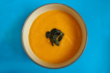 a close-up cream soup of pumpkin and carrot with pumpkin seeds stands in a round gray plate on a blue background top view . gluten-free homemade food