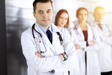 Fototapeta premium Group of professional doctors, standing as a team with crossed arms in a sunny hospital office, ready to help their patients. Medical help, insurance in health care and medicine concept