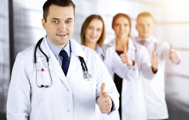 Group of professional doctors are standing as a team with thumbs up in a sunny hospital office, ready to help their patients. Best medical help, insurance in health care and medicine concept