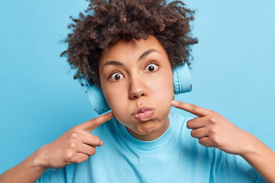 Close up shot of young African American woman pouts cheeks blows face has stunned expression dressed in casual clothes wears stereo headphones listens audio track isolated on blue background