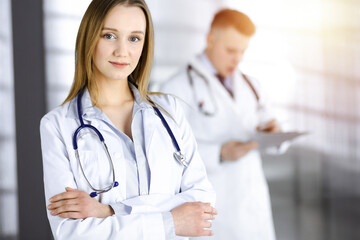 Professional beautiful woman-doctor with a stethoscope is standing with crossed arms in a sunny clinic. Young doctors at work in a hospital. Modern medicine and healthcare concept