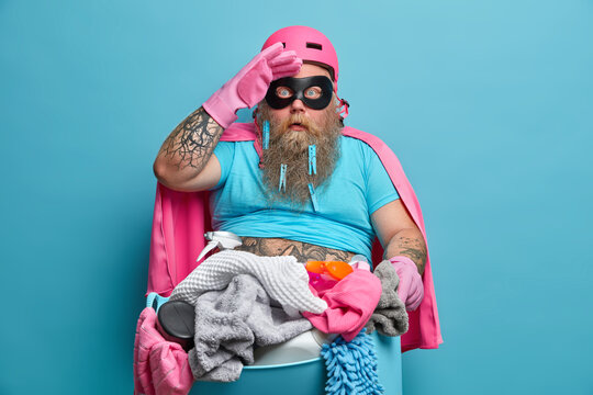 Surprised bearded man stares at camera keeps hand on forehead poses with basin of laundry busy doing household chores wears superhero costume isolated over blue background. Housekeeping concept