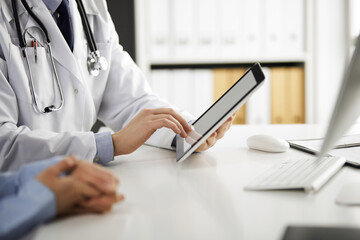 Unknown male doctor and patient woman discussing current health examination while sitting in clinic and using tablet computer, closeup of hands. Medicine and healthcare concept