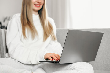 A young blonde woman is sitting on the couch and using her laptop. Distance education. Work online. Freelancer girl