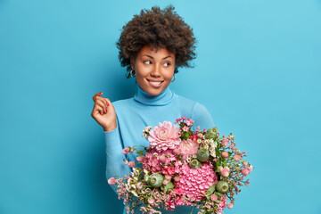 Adorable charming young African American woman gets flowers as birthday gift looks thoughtfully aside smiles gently dressed in poloneck isolated over blue background. Women and holiday concept