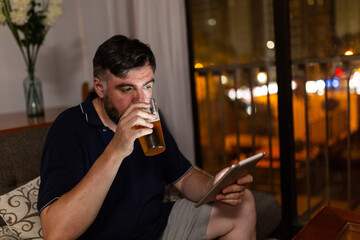 bearded man on sofa uses digital tablet and drink a beer in dining room, night