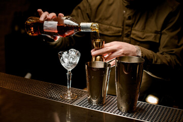 steel shaker cups and glass with ice cubes stand on bar counter and man bartender pours drink