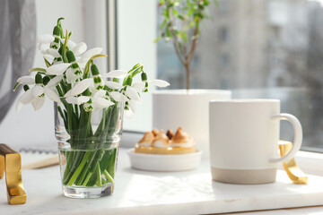Fototapeta na wymiar Beautiful snowdrops, cup of drink and dessert on white tray near window. Space for text