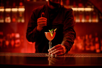 man bartender holds with his hand glass with bright alcoholic cocktail on bar counter.