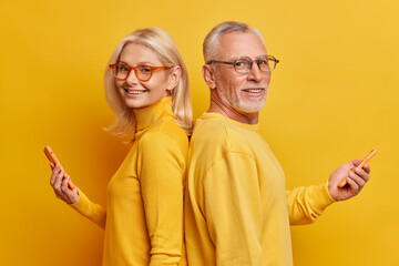 Photo of cheerful senior woman and man stand in profile with modern gadgets hold smartphone have technology addiction wear casual clothes isolated over yellow background. Pensioners use devices