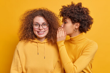 Two best friends gossip share secrets with each other. Afro American woman whispers gossips into companions ear who has surprised cheerful expression excited by latest news. Confidentiality concept