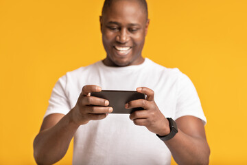 Smiling adult african american male in white t-shirt looks in phone and rejoices to victory