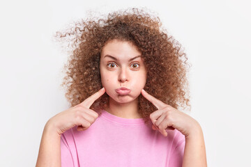 Funny curly haired European woman blows cheeks has fun never looses sense of humor holds breath makes grimace dressed in basic t shirt isolated over white background. People emotions concept - obrazy, fototapety, plakaty