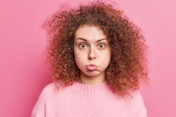 Offended curly haired young European woman blows cheeks makes grimace dressed in casual jumper holds breath looks surprisingly at camera isolated over pink background. Face expressions concept
