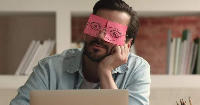 Close up lazy office employee sleeps sit at workplace covering his eyes with sticky notes, open eyes drawn on adhesive papers behind glasses. Overworked, chronic fatigue, no motivated worker concept