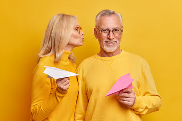 Pleased beatded grey haired man gets kiss from wife pose next to each other hold handmade paper planes isolated over yellow background. Elderly couple feel love in their old age. Relationship