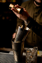 Close-up view of steel shaker cup into which male bartender pours drink from jigger