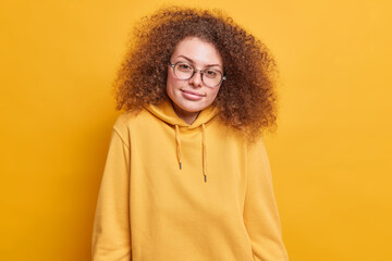 Pleased curly haired pretty female student with serious expression wears transprarent glasses and casual sweatshirt isolated over yellow studio background. People human face expressions concept