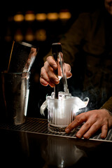 Fototapeta na wymiar magnificent view of illuminated steaming glass in which bartender put piece of ice using tongs