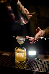 man at bar gently pours cocktail from shaker cup into old-fashioned glass through sieve