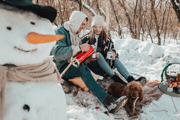 Beautiful couple in love on a winter picnic, drinking tea and smiling, making a snowman