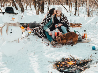 Beautiful couple in love on a winter picnic, drinking tea and smiling, playing with the dog near the fire
