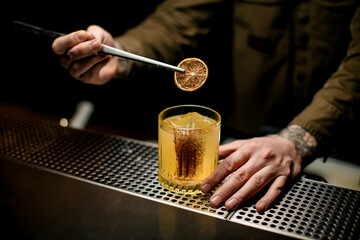 man at bar carefully holds slice of citrus with tweezers over glass with cocktail