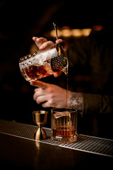 close-up of bartender's hand holds mixing cup and pours cocktail into glass