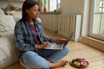 Sleepy young dark skinned woman spending weekend at home sitting barefoot on floor with portable computer on her lap, watching series online via video streaming website, eating sweet snacks