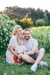 Fototapeta premium Adorable young family with baby boy toddler having weekend in a city park. Blond haired beautiful mom, handsom dad funny kid sitting on grass near blossom roses bush