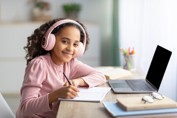 Online education. Smiling teen girl in headset using laptop and taking notes, looking at camera