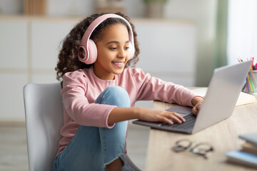 Happy teenage girl chatting on laptop, wearing wireless headphones, relaxing sitting at desk at home