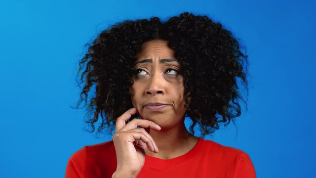 Thinking mixed race woman looking up and around on blue studio background. Pensive face expressions. Pretty afro model with attractive appearance. 