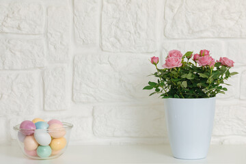 A clear bowl full of pastel colored Easter eggs next to a white wall. Spring flowers in pastel colors. Copy space 