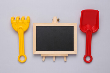 Toy shovel and rake and empty chalk board on gray background. Beach vacation concept. Copy space