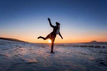 Young girl in winter clothes with a scarf and hat with pompom, balancing on one leg, on frozen plain with shining sun on blue sky in background.