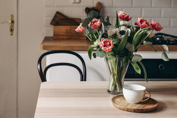 Spring breakfast still life with cup of coffee. Red tulips, white anemone flowers and eucalyptus branches. Glass vase on wooden table. Blurred kitchen background with old chair. Scandinavian interior.