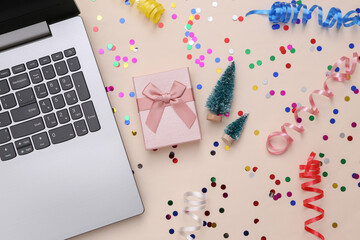 Laptop, Christmas mini trees, gift box and colored serpentine with confetti on beige background. Christmas background