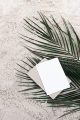 Tropical summer stationery still life. Empty greeting card mock-up, craft envelope on green date palm leaves. Sandy beach background, desert. Flat lay, top view. Exotic island vacation, travel concept