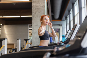 Fototapeta premium Red-haired fitness woman showing thumbs up in modern gym. Happy fit slim woman works out on a treadmill, cardio workout, healthy lifestyle.