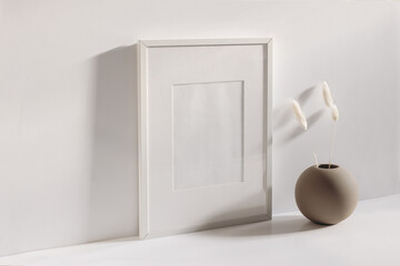 Modern summer stationery still life scene. Beige ball shaped vase with dry lagurus grass in sunlight with long shadows. Empty black vertical picture frame mockup. White table and wall background.