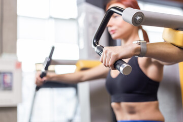 Muscular fit redhead woman trains muscles of shoulders in exercise machine in the gym