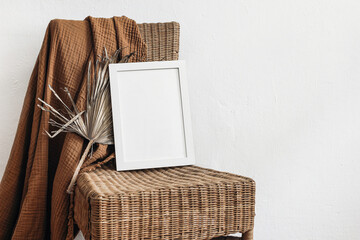 Exotic tropical boho interior still life. Blank white vertical picture frame mock-up on rattan chair. Cinnamon muslin plaid with dry palm leaf. White wall background. Empty copy space, no people.