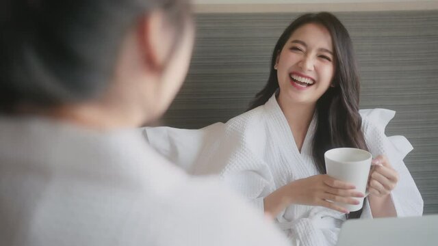 asian female girlfrends in bathrope sit relax hand hold hot coffee cup casual talk with her friend with fun and humor laugh smile happiness joyful on bed in hotel room